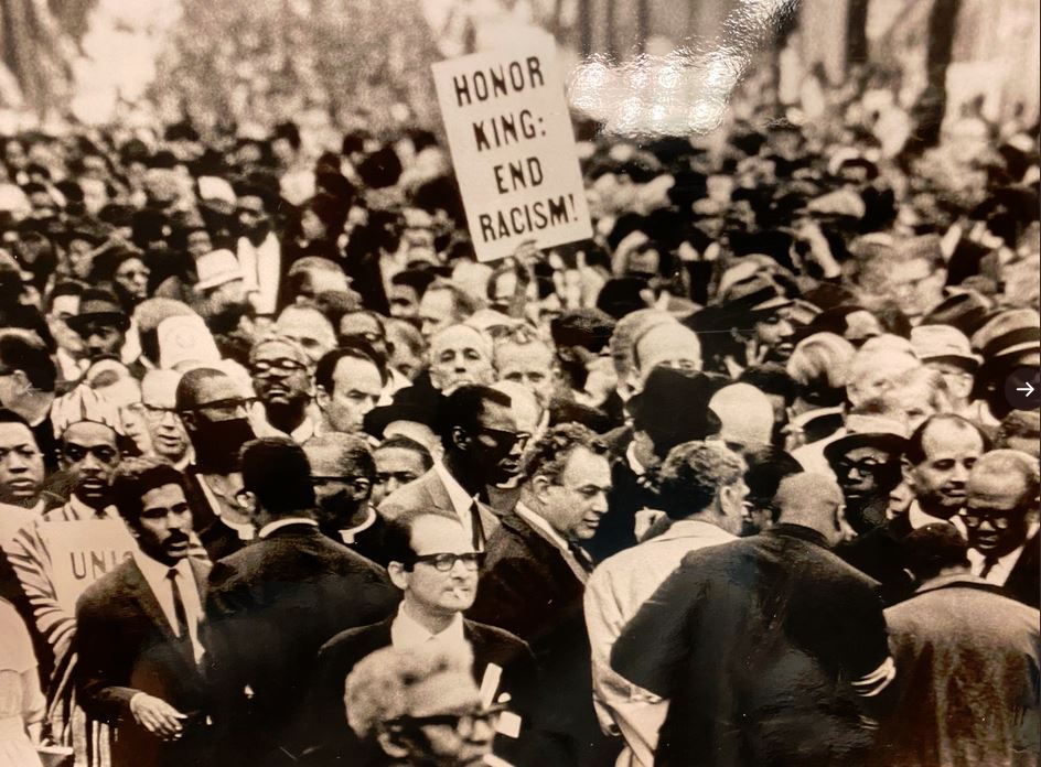 Black and white photo. Crowd of people at 1960s protest. One person holds up sign "Honor King: End Racism!"