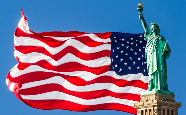 Statue of Liberty with US Flag waving in background against blue sky