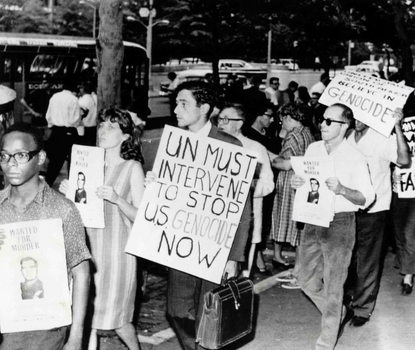 Black and white photograph of protesters holding signs that read, "Wanted for Murder" and "UN Must Intervene to Stop U.S. Genocide Now."