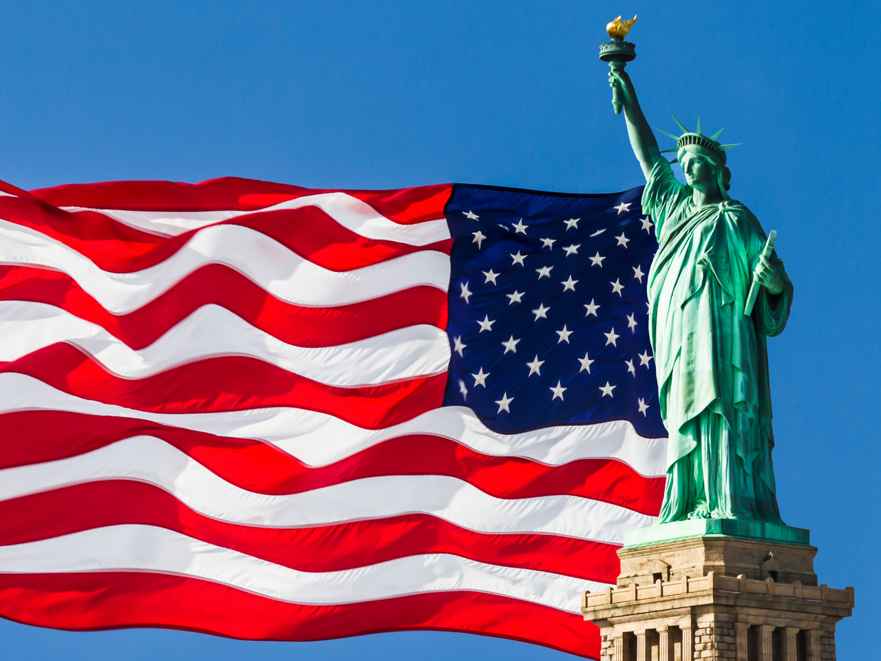 Statue of Liberty with US Flag waving in background against blue sky