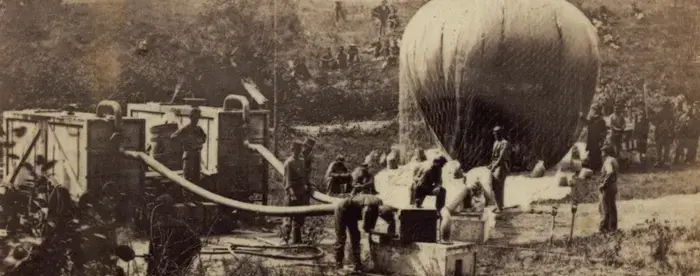 Photograph of five men inflating a hot air balloon. Balloon is partially inflated in the image, and other soldiers can be seen in the background. Image was taken outside, with a hill and trees in the background. 