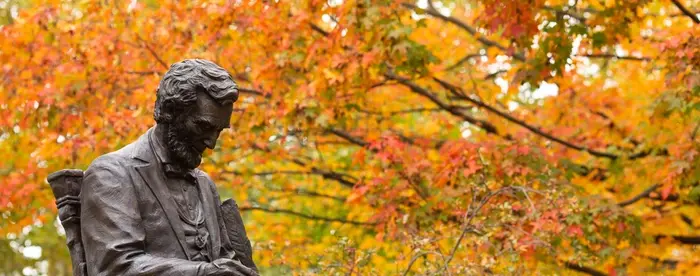 Photograph taken during autumn, showing statue of Lincoln with quill in hand, on Gettysburg campus, fall folliage in background