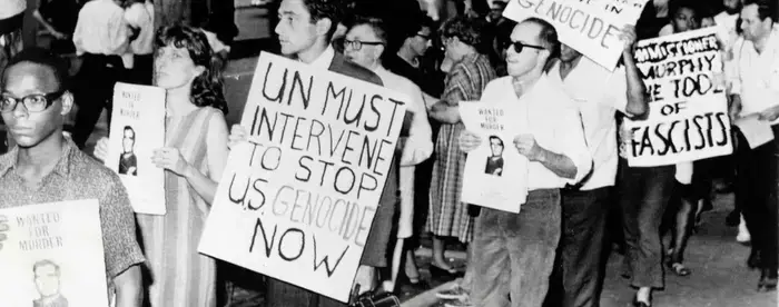 Black and white photograph of protesters holding signs that read, "Wanted for Murder" and "UN Must Intervene to Stop U.S. Genocide Now."