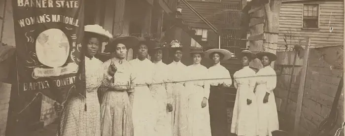 Nine Black women posed standing with one holding a banner that says "Woman's National Baptist Convention"