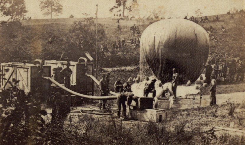Photograph of five men inflating a hot air balloon. Balloon is partially inflated in the image, and other soldiers can be seen in the background. Image was taken outside, with a hill and trees in the background. 