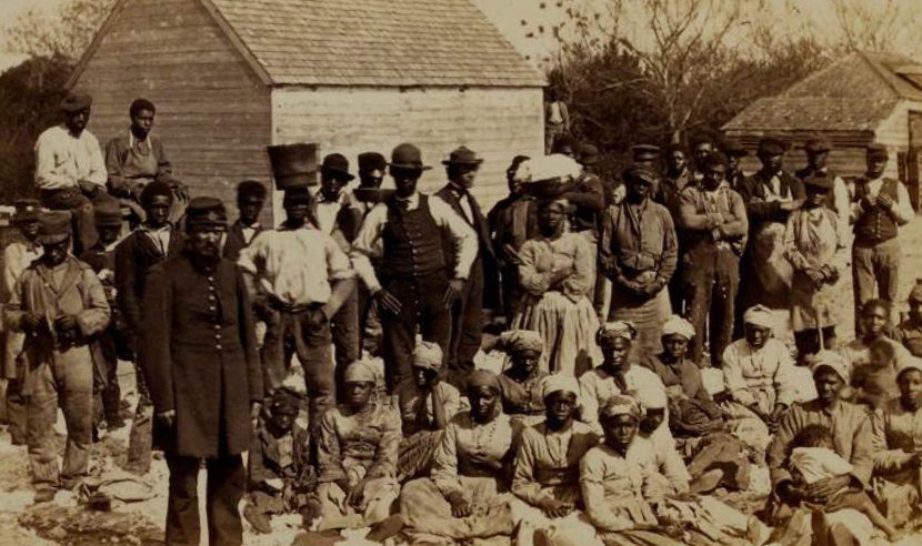 Group of emancipated enslaved people sitting in yard. With buildings visible in background.
