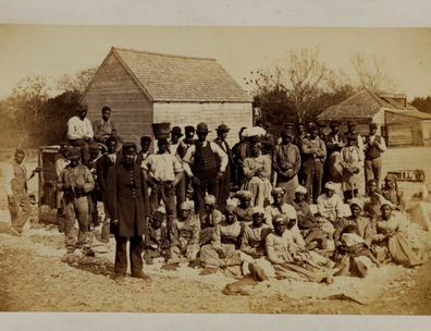 Group of emancipated enslaved people sitting in yard. With buildings visible in background.