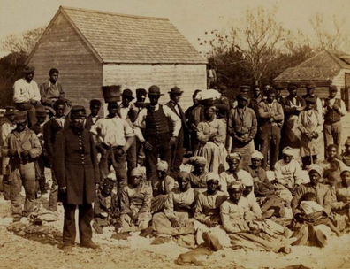 Group of emancipated enslaved people sitting in yard. With buildings visible in background.